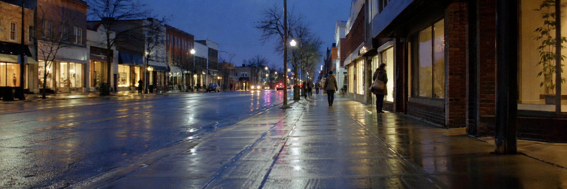 A main street in a small city at dusk, shop windows lit and reflected on wet pavement, a few people walking at a distance